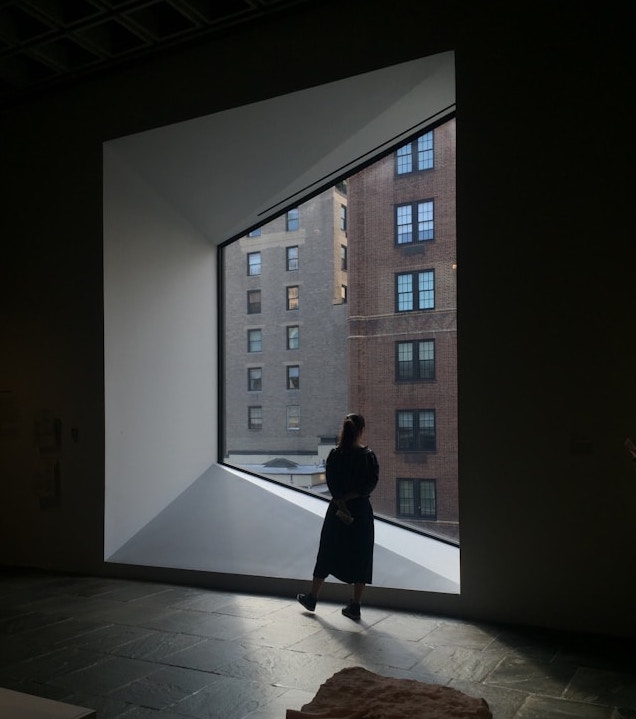 A woman in a dark stone room looking out of a window facing two tall urban apartments